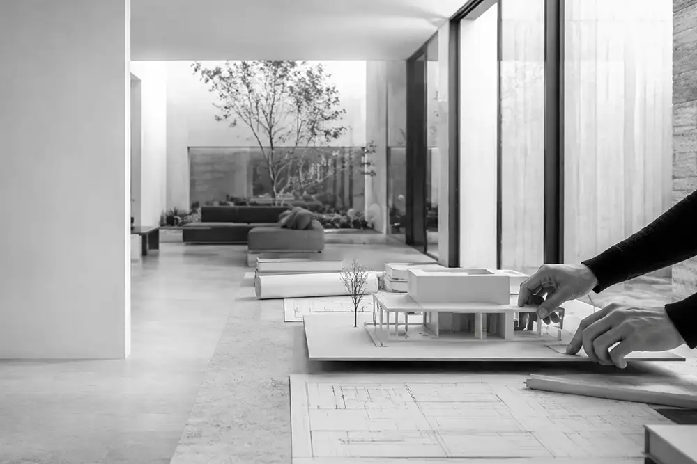 Interior de un estudio de arquitectura con una maqueta y planos sobre una mesa, en blanco y negro.